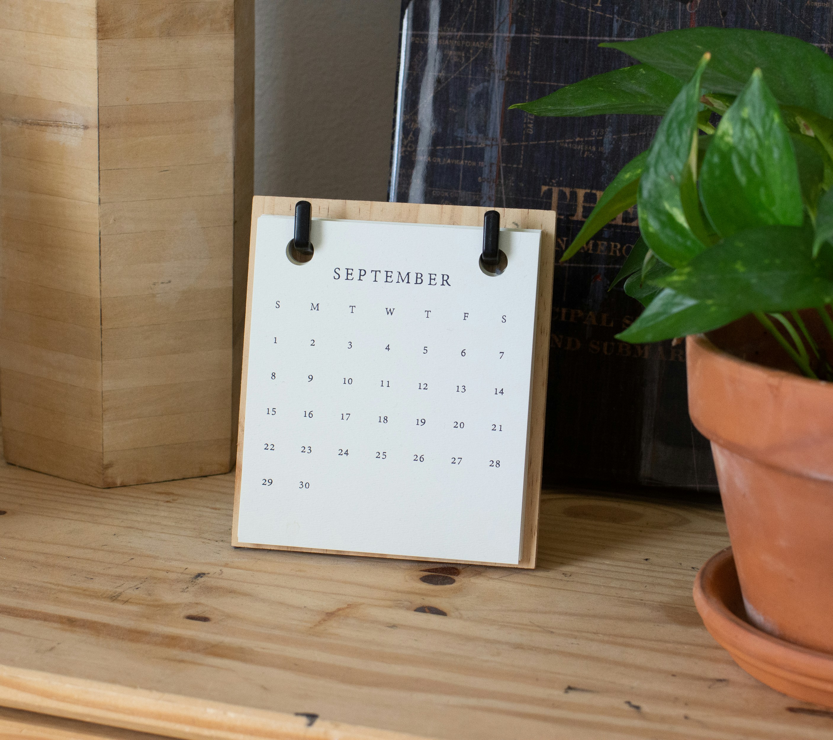 Minimalist desk calendar displaying the month of September, placed on a wooden surface next to a potted green plant and books.